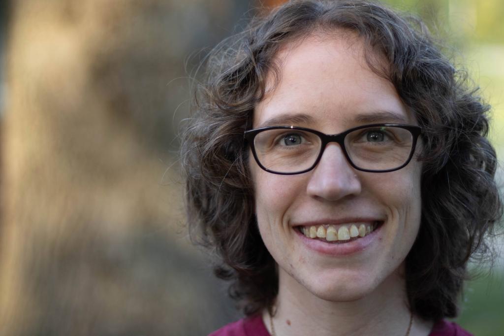white woman with brown curly neck length hair is smiling and wearing black glasses