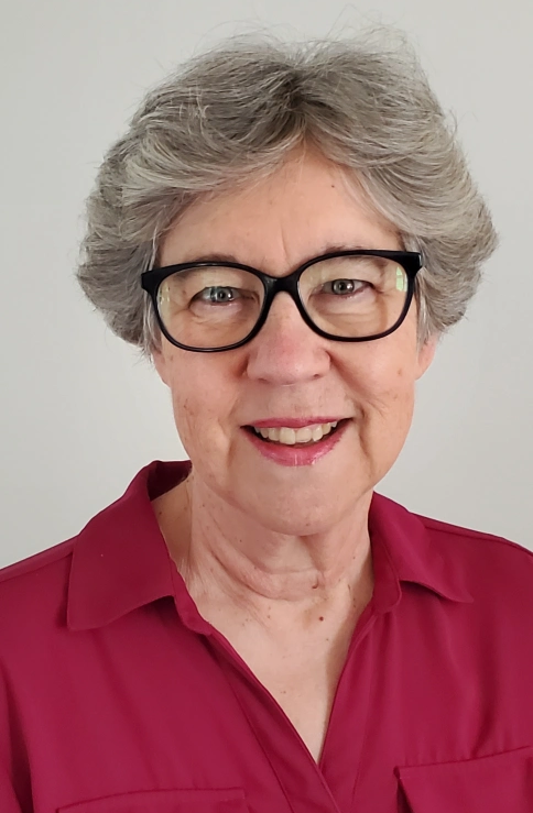 white woman with wispy grey hair is wearing black glasses and deep pink collared shirt.